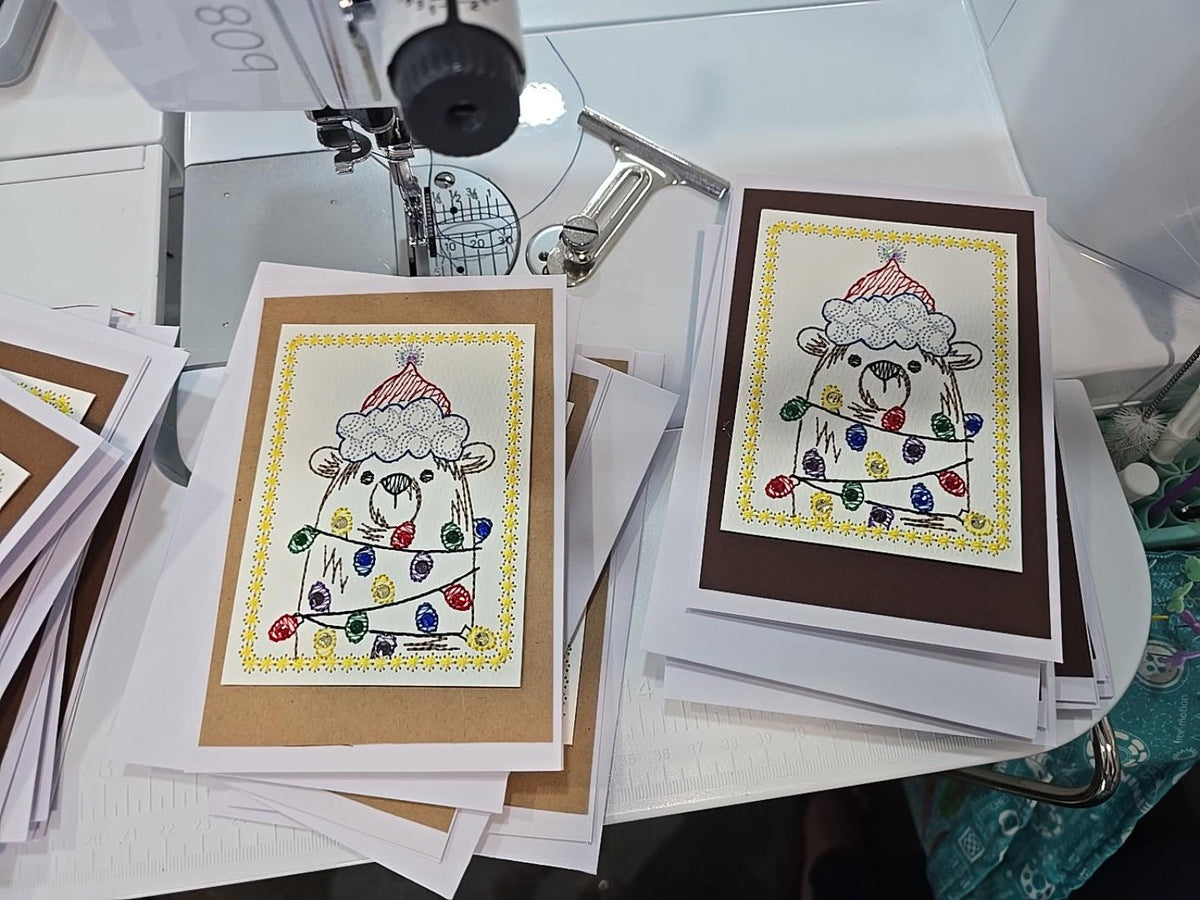 Kindred Carry embroidered Christmas greeting card with a bear in a Santa hat wrapped in colorful lights, available with kraft or dark brown backing