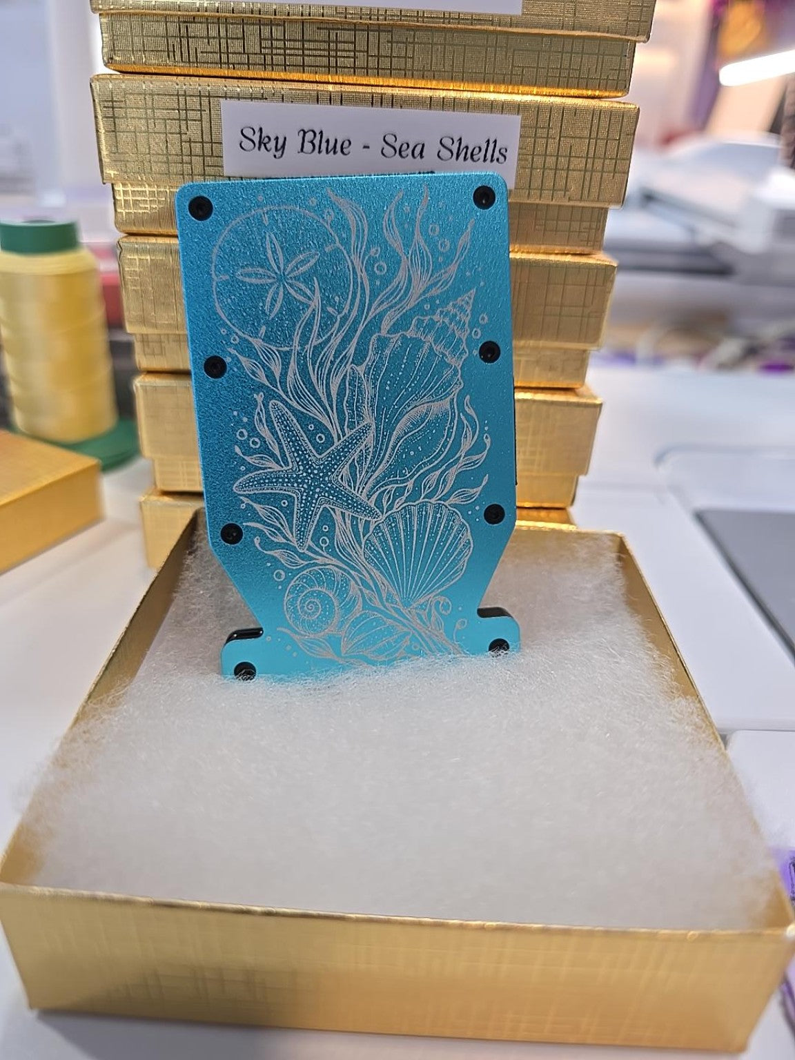 Sky-blue Kindred Carry Notch RFID metal wallet with laser engraved seashells, starfish, sand dollar and flowing sea grass, shown in its gold gift box